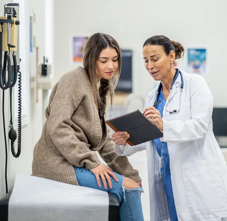 A doctor in a white coat shows a tablet to a patient with long hair wearing a brown sweater in a medical office.